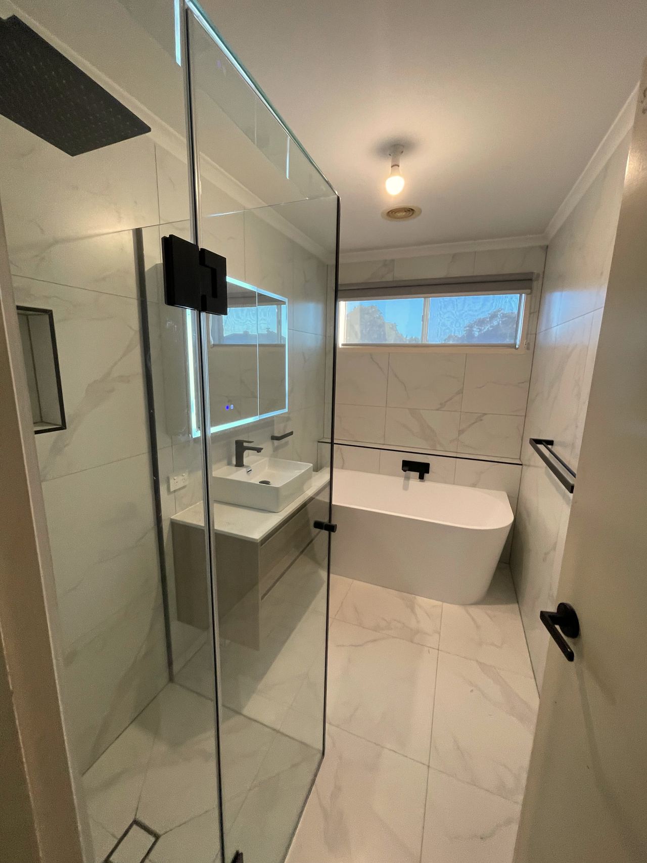 Renovated bathroom in Cranbourne East with a freestanding bath under the window, wall-hung vanity, illuminated mirror cabinet and frameless shower