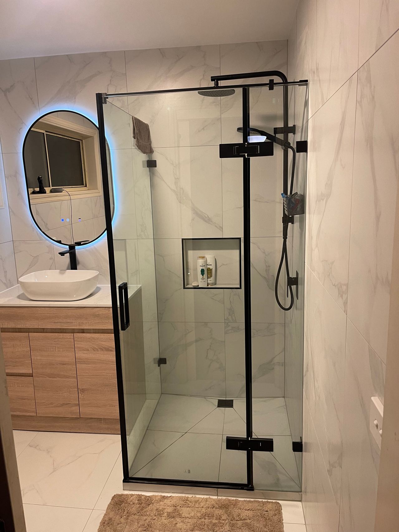After: the same Cranbourne North bathroom with marble-look tiles, black-framed shower and backlit oval mirror